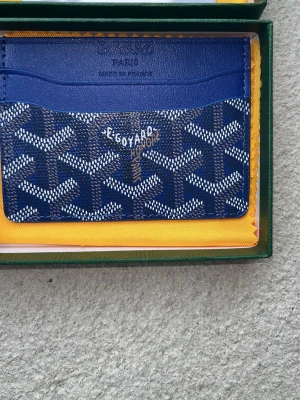Blå plånbok från Goy4ard - Snygg blå korthållare från Goy4ard med klassiskt mönster i vitt och brunt. Tillverkad i exklusivt läder och canvas, har flera fack för kort och är perfekt för dig som vill ha något både stilrent och unikt. Levereras i originalask med gul tygpåse.