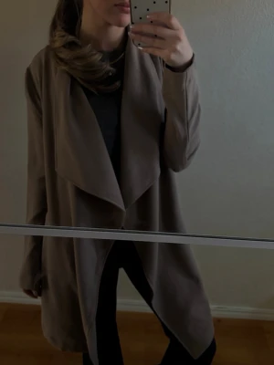 Beige vattenfalls-kappa - Stilren tunn beige/brun kappa med vattenfalls-krage och öppen front. Mellan lång modell med avslappnad passform och mjukt, följsamt tyg. 