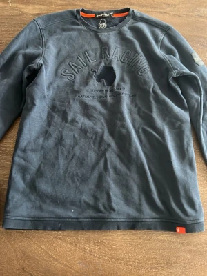 Mörkblå sweatshirt från Sail Racing - Mörkblå sweatshirt från Sail Racing med broderad logotyp och Antartic Expedition-tryck på bröstet. Tröjan har rund hals, långa ärmar och ribbade muddar. Materialet är mjukt och bekvämt, perfekt för en avslappnad stil.
