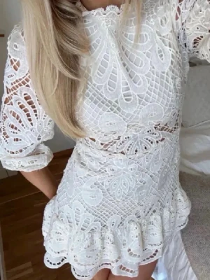 Vit spetsklänning med volang - Supersnygg vit kortklänning i spets med volangkant och genombrutet mönster. Klänningen har långärmade puffärmar, hög hals och markerad midja. Perfekt för studenten😍
