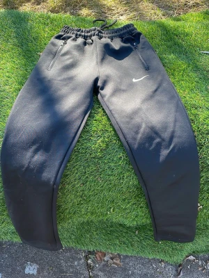 Svarta Nike mjukisbyxor med dragkedjor - Säljer ett par svarta mjukisbyxor från Nike med dragsko i midjan och dragkedjor vid fickorna. Byxorna har vita detaljer och Nike-logga på låret. Tillverkade i ett mjukt och stretchigt material som passar perfekt för chill eller träning.