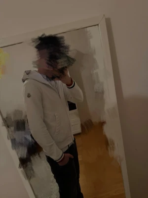 Vit hoodie från Moncler med dragkedja - Säljer en vit hoodie från Moncler med dragkedja framtill och huva. Tröjan har Moncler-logga på ärmen och ribbade muddar med röd, vit och blå rand. Snygg och stilren design som passar till det mesta. Perfekt för dig som gillar exklusiva streetwear-plagg.