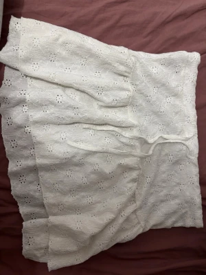 Vit kortkjol med broderade detaljer - Söt vit kortkjol med broderade blommönster och volangkant. Kjolen har knytband i midjan och är gjord i ett lätt bomullstyg som passar perfekt till varma dagar.