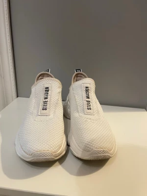 Vita slip-on sneakers Steve Madden - Snygga vita slip-on sneakers från Steve Madden med stickad ovandel och bred sula. Skorna har logotyp på både plösen och hälen, samt en sportig och modern design. Perfekta för dig som gillar stilrena och bekväma sneakers.