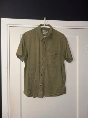 Olivgrön kortärmad skjorta från Solid - Kortärmad skjorta från Solid i olivgrön färg med klassisk krage och bröstficka. Skjortan har knappar framtill och är tillverkad i ett lätt, strukturerat material som passar perfekt till varmare dagar. Regular fit för en avslappnad look.