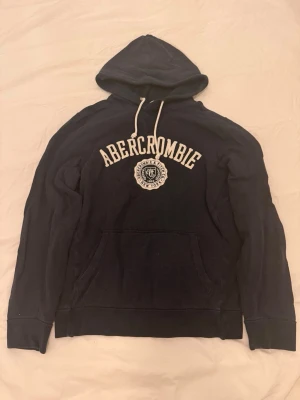 Mörkblå hoodie från Abercrombie - Snygg mörkblå hoodie från Abercrombie med vit broderad logga och text på bröstet. Klassisk känguruficka framtill och justerbar huva med vita snören. Perfekt för en avslappnad och trendig stil.