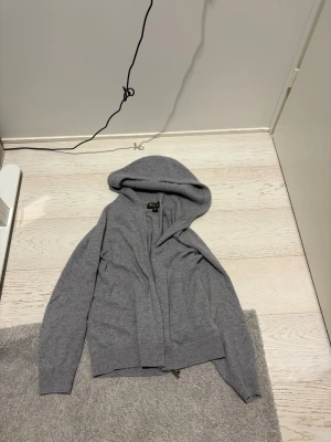 Ljusgrå hoodie i kashmir från Loro Piana - Exklusiv ljusgrå hoodie från Loro Piana i 100% kashmir. Tröjan har dragkedja framtill, ribbade muddar och en stor huva. Perfekt för dig som gillar lyxiga och mjuka material. Tillverkad i Italian. Size är s eller xs