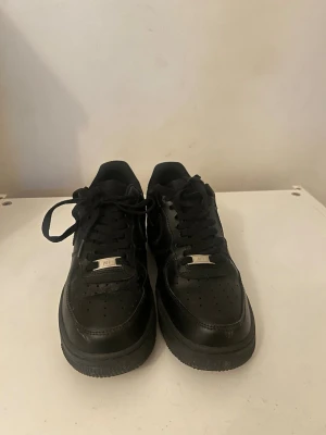 Nike Air Force 1 helsvarta sneakers - Säljer ett par klassiska Nike Air Force 1 sneakers i helsvart läder. Skorna har lågt skaft, perforerad tåbox och ikonisk Swoosh på sidorna. Perfekta för en clean och stilren look. Metall-detalj med 'AF1' på snörningen.