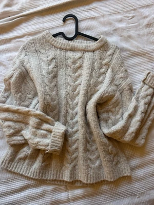 Beige kabelstickad tröja - Mysig beige kabelstickad tröja med breda flätmönster framtill och på ärmarna. Tröjan har rund halsringning, långa ärmar och ribbade muddar vid ärmslut och nederkant. Perfekt för kalla dagar och enkel att matcha med jeans eller kjol.