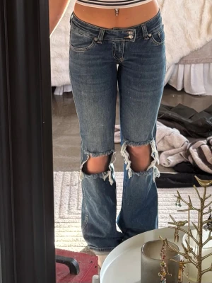 Blå slitna jeans med hål vid knäna - blå jeans med raka ben och stora slitna hål vid båda knäna. Jeansen har låg midja.