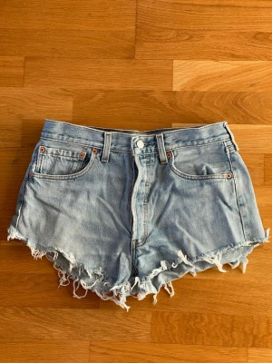 Levi's 501 ljusblå jeansshorts med fransar i storlek W33 - Säljer ett par klassiska ljusblå jeansshorts från Levi's 501 med råa, fransiga kanter och femficksmodell. Shortsen har hög midja och är tillverkade i bomull. Perfekta för sommaren och ger en avslappnad vibe. Storlek W33.
