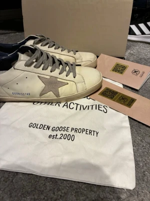Golden Goose vita sneakers med stjärna - Golden Goose sneakers i vitt läder med grå mockastjärna på sidan och grå skosnören. Hälpartiet är mörkblått och skon har GGDB/SSTAR-tryck i blått. Klassisk låg modell med slitna detaljer för en cool look.