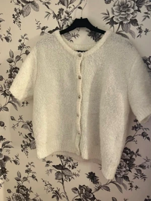 Vit kortärmad kofta med knappar - Mysig vit kofta med korta ärmar och knäppning framtill. Tillverkad i ett fluffigt, mjukt material som ger en avslappnad och trendig look. Perfekt att slänga över en topp för en chill vibe.