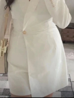 Vit blazerklänning med knapp jumpsuit  - Stilren vit kortklänning i blazer-modell med lång ärm och markerad midja. Klänningen har en guldfärgad knapp framtill och klassisk krage. Materialet känns mjukt och följsamt, perfekt för en chic look.jumpsuit 