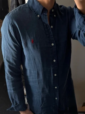 Mörkblå linneskjorta från Ralph Lauren - Klassisk mörkblå linneskjorta från Ralph Lauren med button-down krage och vita knappar. Skjortan har lång ärm och den ikoniska röda broderade loggan på bröstet. Tillverkad i 100% linne. Ganska urtvättad därav priset, men annars bra skick