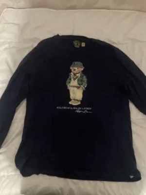 Mörkblå långärmad Polo Ralph Lauren - Snygg mörkblå långärmad tröja från Polo Ralph Lauren med ikoniskt björnmotiv på bröstet. Tröjan har rund halsringning och tryckt logga under motivet. Perfekt för dig som gillar streetwear och klassisk stil. 