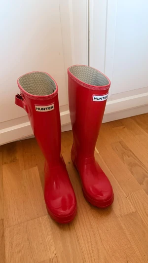 Röda höga gummistövlar från Hunter - Snygga röda gummistövlar från Hunter med klassisk hög modell och justerbart spänne vid skaftet. Andvända ett fåtal gånger men inga tecken på andväning. Pris kan diskuteras! Passar mig som har 36/37