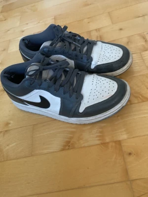 Nike Air Jordan 1 Low grå/vit - Säljer ett par Nike Air Jordan 1 Low sneakers i grått och vitt med svart swoosh. Skorna har klassisk låg siluett, perforerad tåbox och snörning. Ovandelen är i läder och textil, vilket ger både stil och komfort.