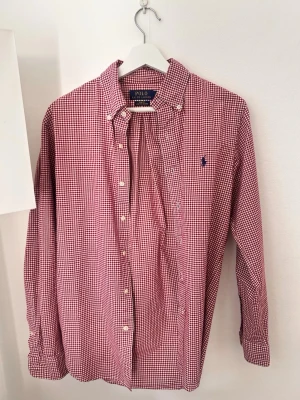 Rutig skjorta från Polo Ralph Lauren - Klassisk röd och vit rutig skjorta från Polo Ralph Lauren med button-down krage och broderad logga på bröstet. Skjortan har långa ärmar, knappar framtill och är tillverkad i mjuk bomull. Perfekt för en stilren och avslappnad look.