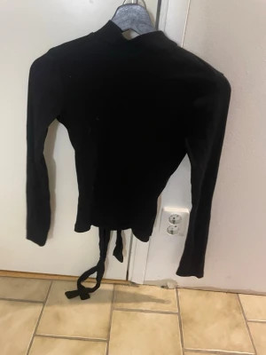 Svart omlottopp med knytband - Säljer en svart omlottopp med långa ärmar och knytband i midjan. Toppen har en hög hals och är figurnära i modellen, perfekt för att skapa en snygg siluett. Materialet känns mjukt och stretchigt, vilket gör den bekväm att bära hela dagen.