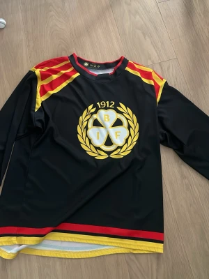 Brynäs IF hockeytröja svart XL - Säljer en svart Brynäs IF hockeytröja för säsongen 24/25 med klubbens klassiska logga i gult och vitt på bröstet. Tröjan har röda och gula detaljer på axlar och nederkant, samt lång ärm. Tillverkad i polyester, perfekt för match eller supporterhäng.