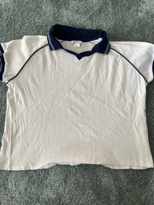 Vit t-shirt med marinblå krage Monki - Vit t-shirt från Monki med marinblå krage och detaljer vid ärmslut. T-shirten har korta ärmar och subtilt randigt mönster i tyget. Perfekt för en sportig och avslappnad look.