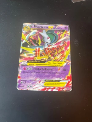 Pokémonkort M Gardevoir EX - Säljer ett samlarkort från Pokémon, M Gardevoir EX med 210 HP. Kortet har en färgstark design med lila, rött och gult samt holografiska effekter. Text på både engelska och japanska. Perfekt för dig som gillar Pokémon och vill utöka din samling.