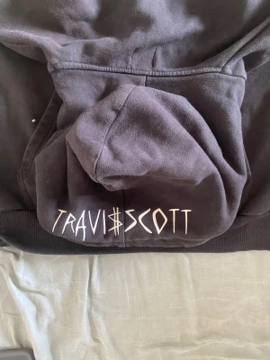  Travis Scott hoodie (real) - Säljer en svart hoodie från Travis Scott med stort vitt smiley-tryck på bröstet och Travis Scott-logga på huvan. Ärmarna har vita detaljer och muddar. Klassisk känguruficka framtill och huva med dragsko. Perfekt för dig som gillar streetwear och unik design.