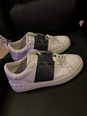 Vita Valentino sneakers med mörkblå detalj - Snygga vita sneakers från Valentino med en bred mörk blå läderdetalj över snörningen. Skorna har klassisk låg profil, vita snören och subtila nitar på hälen. Tillverkade i slätt läder för en clean och exklusiv look. Notera att de är använda men tvättade!