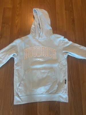 Ljusblå hoodie från Hoodrich - Snygg ljusblå hoodie från Hoodrich med stor magficka och tryck både fram och bak. På framsidan står det 'HOODRICH' och på baksidan 'PRODUCT OF THE STREETS'. Hoodien har huva och ribbade muddar. Perfekt för en avslappnad streetwear-look.