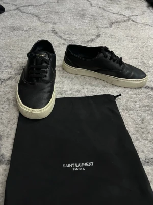 Svarta Saint Laurent lädersneakers - Svarta sneakers från Saint Laurent i mjukt läder med vita, slitstarka gummisulor. Klassisk låg modell med svarta skosnören och diskret logga på hälen. Snygg och stilren design som passar till det mesta. Medföljer original dustbag.