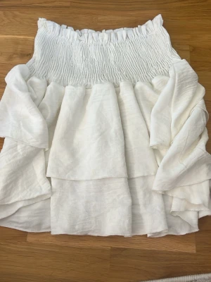 Vit volang tubtopp från SHEIN - Söt vit kjol från SHEIN med bred smock upptill och flera lager volanger.  Kjolen har en luftig, flowy känsla som passar perfekt till sommaren. Tillverkad i ett lätt syntetmaterial. 