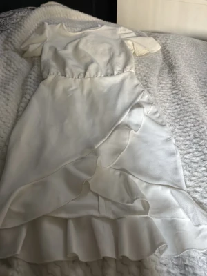 Vit omlottklänning med volang - Säljer en vit kortklänning från bubbleroom med omlottdesign och volangdetaljer på kjolen. Klänningen har korta ärmar och rund halsringning. Perfekt för sommarens alla tillfällen eller skolavslutning ! Materialet känns mjukt och har en lätt glans.