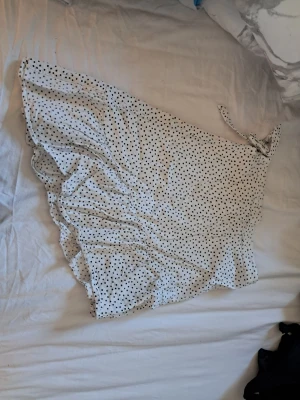 Vit omlottkjol med svarta prickar - Söt vit omlottkjol med svarta prickar och volangkant. Kjolen har knyt i midjan och är kort i modellen, ganska genomskinlig, perfekt för varma dagar. Lätt och luftig känsla, passar till många olika stilar.