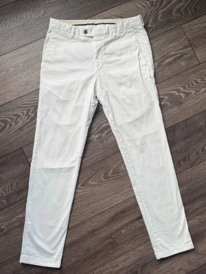 Vita chinos  - Snygga vita chinos från Oscar Jacobson. Byxorna har sidofickor, bälteshällor och två bakfickor. Perfekta för en clean och stilren look. Tillverkade i ett mjukt och lätt material som känns bekvämt hela dagen. Där är en liten svart fläck där bak på benet och och en annan fläck där fram som knappt syns, man ser de på sista bilden. 