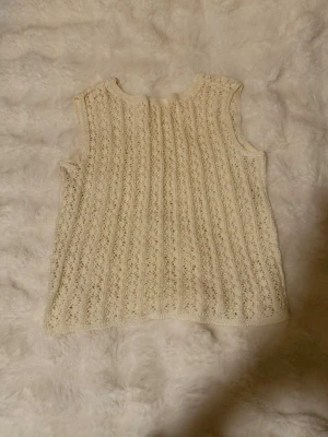 Stickad ärmlös väst i offwhite - Säljer en stickad ärmlös väst i offwhite med ribbade detaljer och hålmönster. Perfekt att styla över en skjorta eller t-shirt för en trendig lager-på-lager-look. Materialet känns mjukt och följsamt.