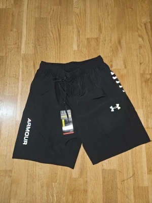 Svarta träningsshorts från Under Armour - Snygga svarta träningsshorts från Under Armour med logga på benet och texten 'ARMOUR' längs sidan. Shortsen har elastisk midja med snörning och är tillverkade i ett lätt och snabbtorkande syntetmaterial. Perfekta för gymmet eller löprundan.