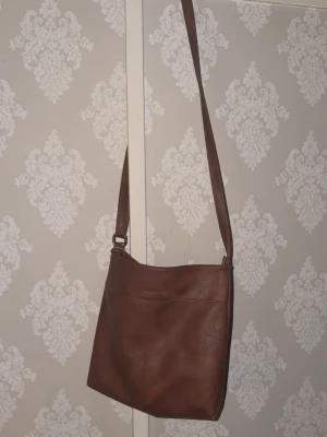 Brun axelväska i läder - Snygg brun axelväska i läder med enkel, stilren design och justerbar axelrem. Väskan har en fyrkantig form och en diskret söm framtill. Verkligen tidlös och perfekt nu till våren!