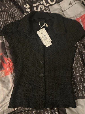 Svart kortärmad blus från Gina   - Trendig svart blus från Gina  med korta ärmar och krage. Blusen har en smickrande figurnära passform, knappar framtill och är gjord i ett stretchigt, krinklat tyg som ger en cool struktur. Perfekt att styla med jeans eller kjol.