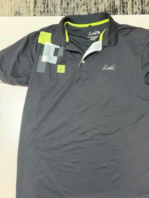 Arnold Palmer mörkblå piké med tryck - Snygg mörkblå pikétröja från Arnold Palmer med grafiskt tryck i vitt, grått och lime på axeln. Pikén har klassisk krage med knappslå och kontrastfärgad insida i lime. Perfekt för dig som gillar sportig stil.