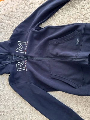 Marinblå hoodie  - Snygg marinblå hoodie med dragkedja framtill och stora RM-bokstäver på bröstet. Hoodien har fickor på magen och justerbar huva med snörning. Perfekt för en chill och stilren look.
