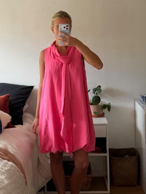 Rosa puffig klänning  - Säljer en söt rosa kortklänning med knyt i halsen. Klänningen är ärmlös och har en lös passform som ger en avslappnad och trendig look. Perfekt för fina tillfällen och  enkel att styla med accessoarer. Storleken är L men fassar för S/M 