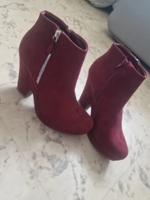 Vinröda stövletter med klack - Snygga vinröda stövletter med hög klack och dragkedja på sidan. Skorna har en mjuk mockaliknande yta och rundad tå. Perfekta för dig som vill sticka ut med färg och stil. Passar till både jeans och klänning. Nästan aldrig använda! Stoeltk 38