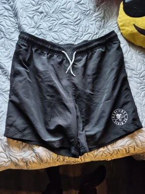 Svarta shorts med snörning - Svarta shorts från Faction med vit snörning i midjan och tryckt logga på vänster ben. De har elastisk midja och är tillverkade i ett lätt syntetmaterial, perfekta för sommarens aktiviteter.