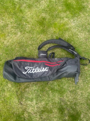 Titleist Premium carrybag - Snygg svart, röd, vit carry golfbag från Titleist! Den är i strålande skick, endast använd några gånger vid rangen, Aldrig spelat runda med den.   Det är en carry bag som har två små stödben som gör att klubborna inte behöver nudda marken. Den har smarta rämmar som används för att ha som ryggsäck, samt vattenflaskhållare och ett flertal smarta fack till övrigt tillbehör! Köptes för 1500kr tidigare. Hör av er vid frågor!🔥🤝