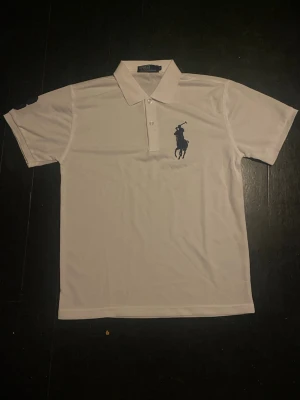 Vit pikétröja från Polo Ralph Lauren - M - polyester - Vit pikétröja från Polo Ralph Lauren med klassisk krage och knappar framtill. Stor mörkblå logga på bröstet. Perfekt för en clean och stilren look. Skön och luftig passform. Polyester