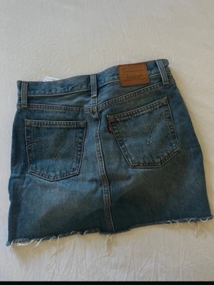Levi's jeanskjol med rå kant - Säljer en klassisk blå jeanskjol från Levi's med rå, fransig kant nertill. Kjolen har fem fickor, knappgylf och bälteshällor. Perfekt för dig som gillar en avslappnad och trendig denimlook.
