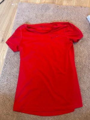 Röd Nike Dri-FIT t-shirt - Röd sportig t-shirt från Nike med Dri-FIT-teknologi. T-shirten har rund halsringning, korta ärmar och en diskret Nike-logga på bröstet. Perfekt för träning eller sportaktiviteter. Materialet är lätt och andas bra.