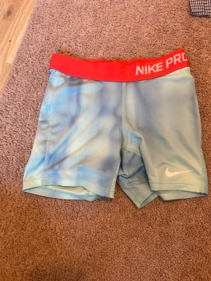 Nike Pro ljusblå träningstights - Säljer ett par Nike Pro träningstights i ljusblått med bred röd resår i midjan där det står 'NIKE PRO' i vitt. Tightsen har en tajt passform och är tillverkade i ett stretchigt funktionsmaterial, perfekt för träning eller sport.