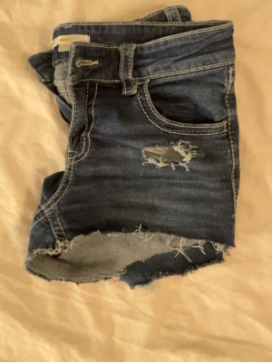 Mörkblå jeansshorts med slitningar - Säljer ett par mörkblå jeansshorts med rå kant och slitningar framtill. Shortsen har klassisk femficksmodell, synliga sömmar i kontrastfärg och en snygg detalj med sliten look. Perfekta för en avslappnad och trendig stil.
